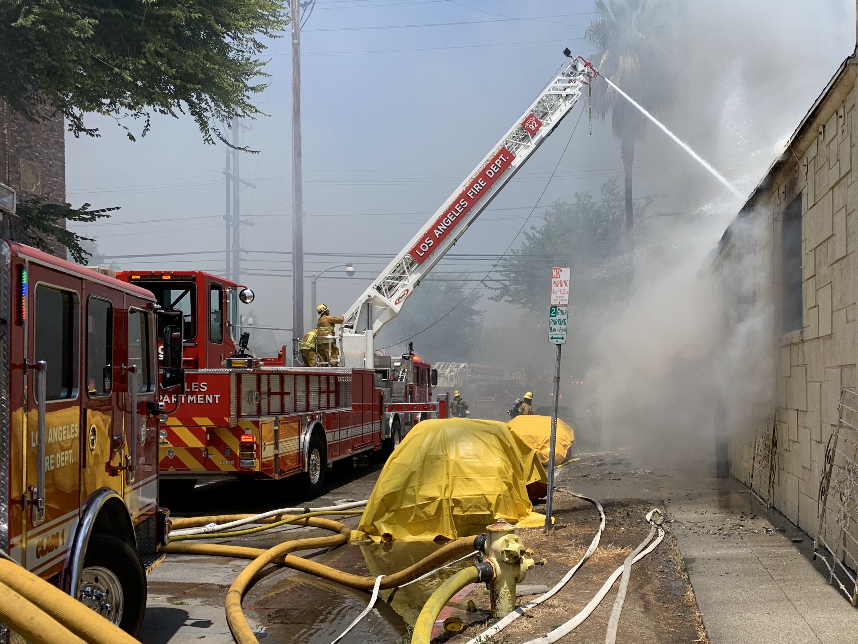 Two cars, draped with yellow tarps, are shown in front of fire trucks with equipment in use around them